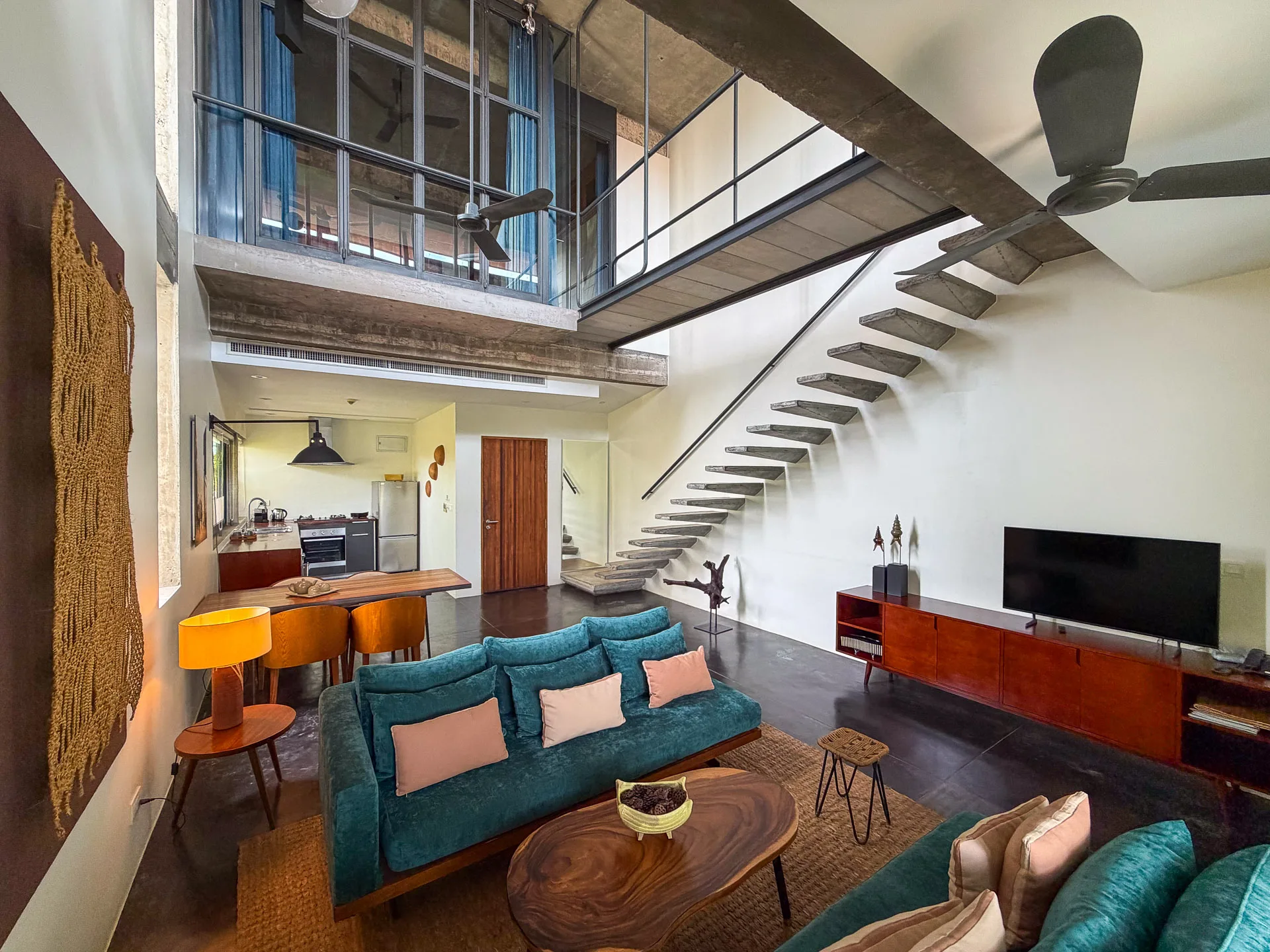Double-height loft living room with teal sofas, floating concrete stairs and a mezzanine bedroom at Urban Residences